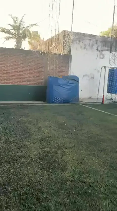 Cancha abierta de césped sintético con arco y tanque de agua azul junto al muro