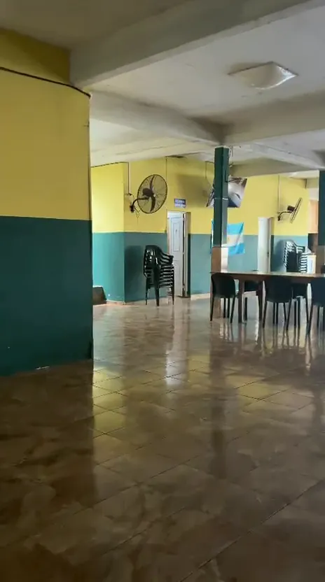 Salón con paredes verdes y amarillas, ventiladores de pared y mesas largas